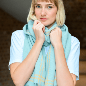 Woman wearing a soft pastel houndstooth lambswool scarf in shades of mint and yellow styled with a white t-shirt. She stands indoors with a calm, happy expression. The scarf features a visible "Just Like Red" label.