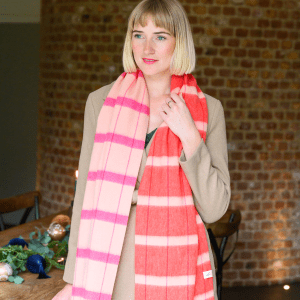 Woman wearing a soft pastel checked lambswool scarf in shades of beige, pink and vibrant red styled with a green dress and a beige coat. She stands indoors with a calm, happy expression. The scarf features a visible "Just Like Red" label.