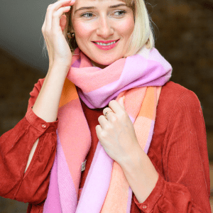 Woman wearing a soft pastel checked lambswool scarf in shades of beige, pink and orange styled with a maroon dress. She stands indoors with a calm, happy expression. The scarf features a visible "Just Like Red" label.