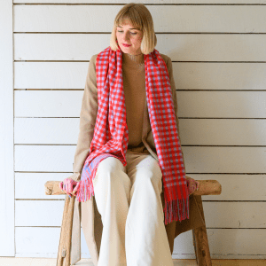 Woman wearing a vibrant gingham check lambswool scarf in shades of red and blue styled with a beige jumper and cream trousers. She i ssitiing indoors with a calm, happy expression. The scarf features a visible "Just Like Red" label.