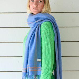 Woman wearing a soft pastel houndstooth lambswool scarf in shades of blue and orange styled with a green jumper. She stands indoors with a calm, relaxed expression. The scarf features a visible "Just Like Red" label.