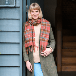 A close-up, high-contrast flat lay photograph of a vibrant red lambswool fabric in a traditional check or tartan pattern. The fabric is folded to show its texture and the design is framed by a bold, solid red border. The text overlay reads, "COLOUR."
