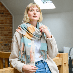 Woman wearing a soft pastel checked lambswool scarf in shades of peach, mint, yellow and beige, styled with a white t-shirt. She stands indoors with a calm, relaxed expression. The scarf features a visible "Just Like Red" label.